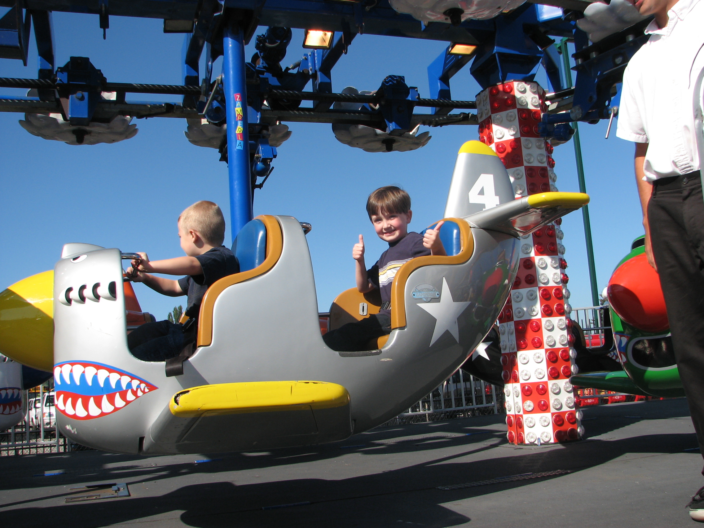 A four year old gives two thumbs up from the backseat of a kiddie carnival jet plane ride.
