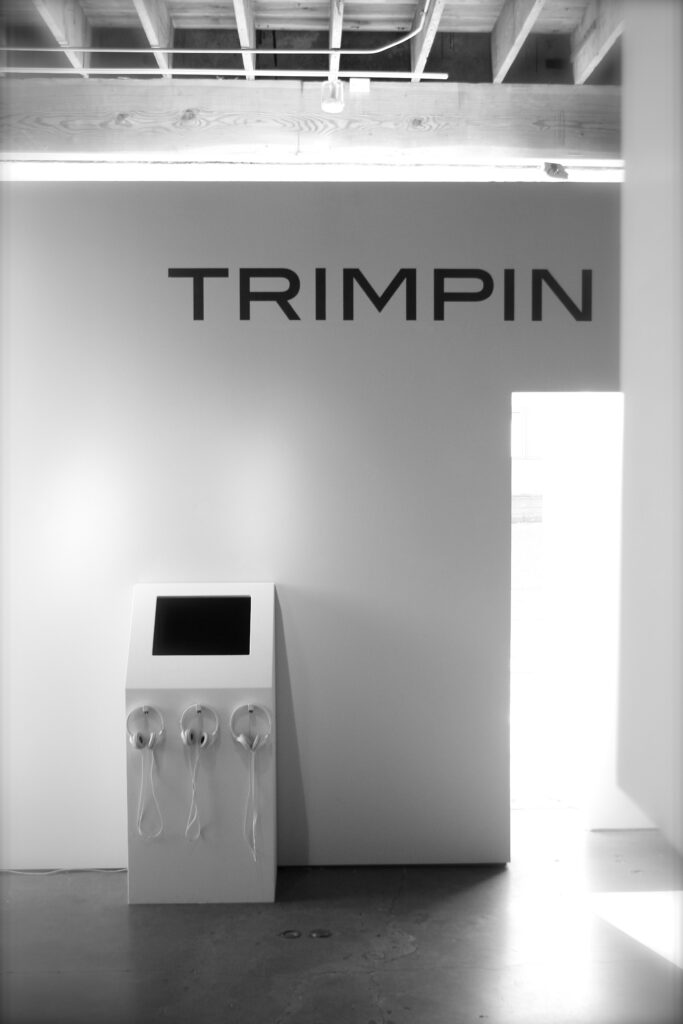 an all white, futuristic entryway features an ATM-like box with headphones and the name Trimpin in all caps at the top of the wall